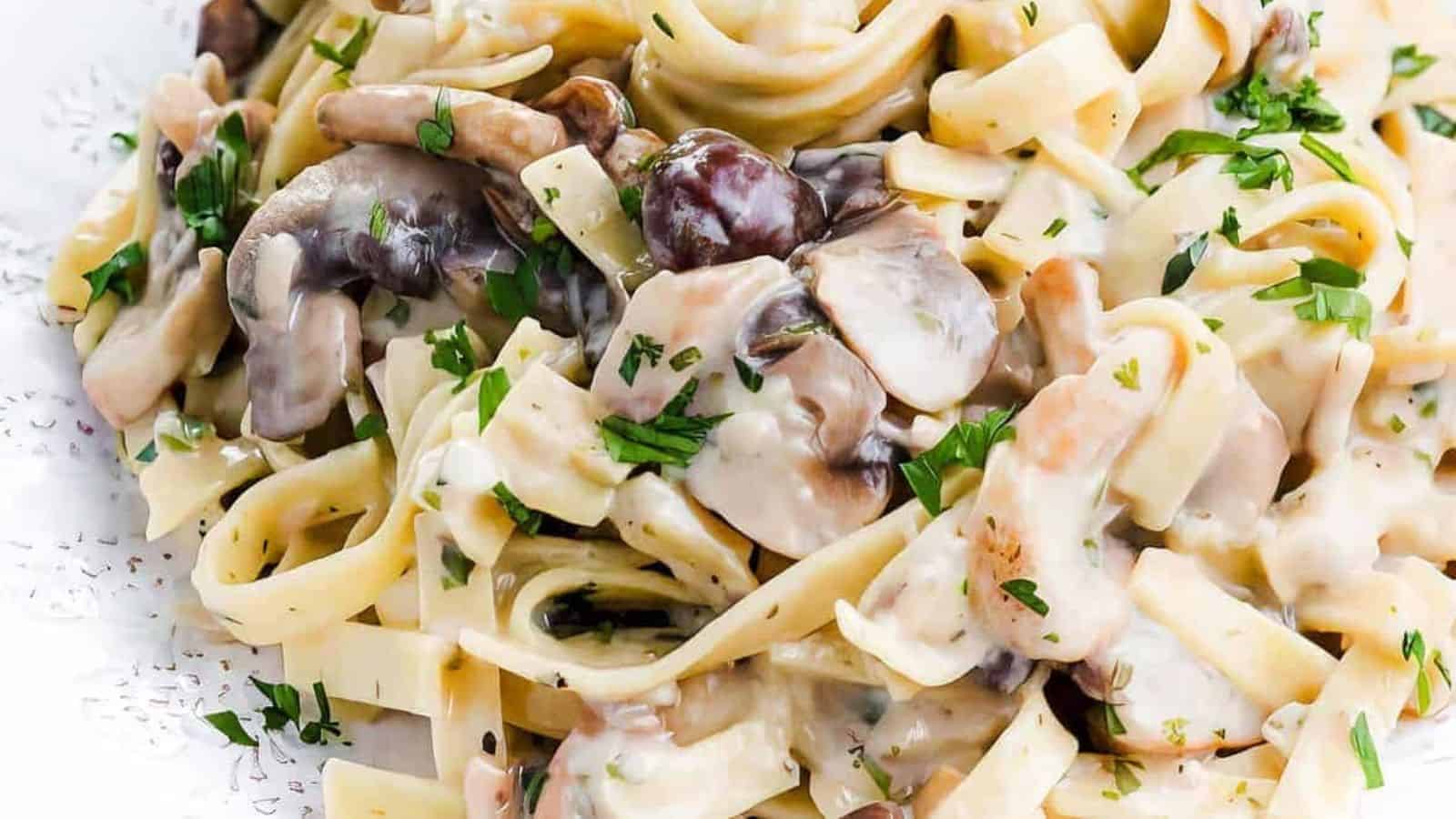 A close-up photo of a white plate overflowing with creamy tagliatelle pasta and sliced cremini mushrooms. A silver fork rests on the side of the plate.