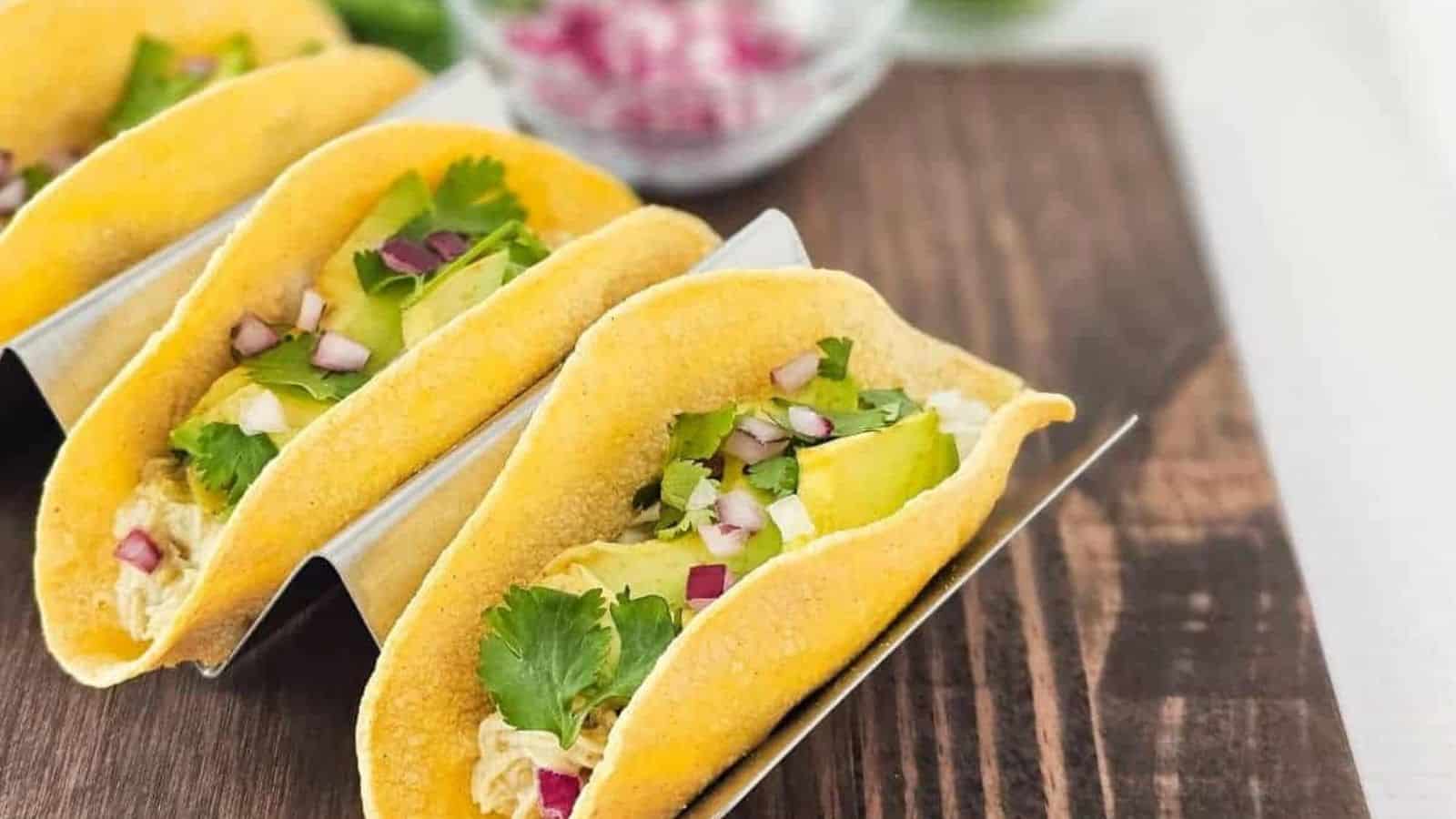 Three chicken tacos topped with cilantro, avocado and red onions on a wooden cutting board.