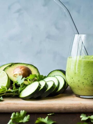 A green smoothie with cucumbers, cilantro and avocado on a wooden cutting board.