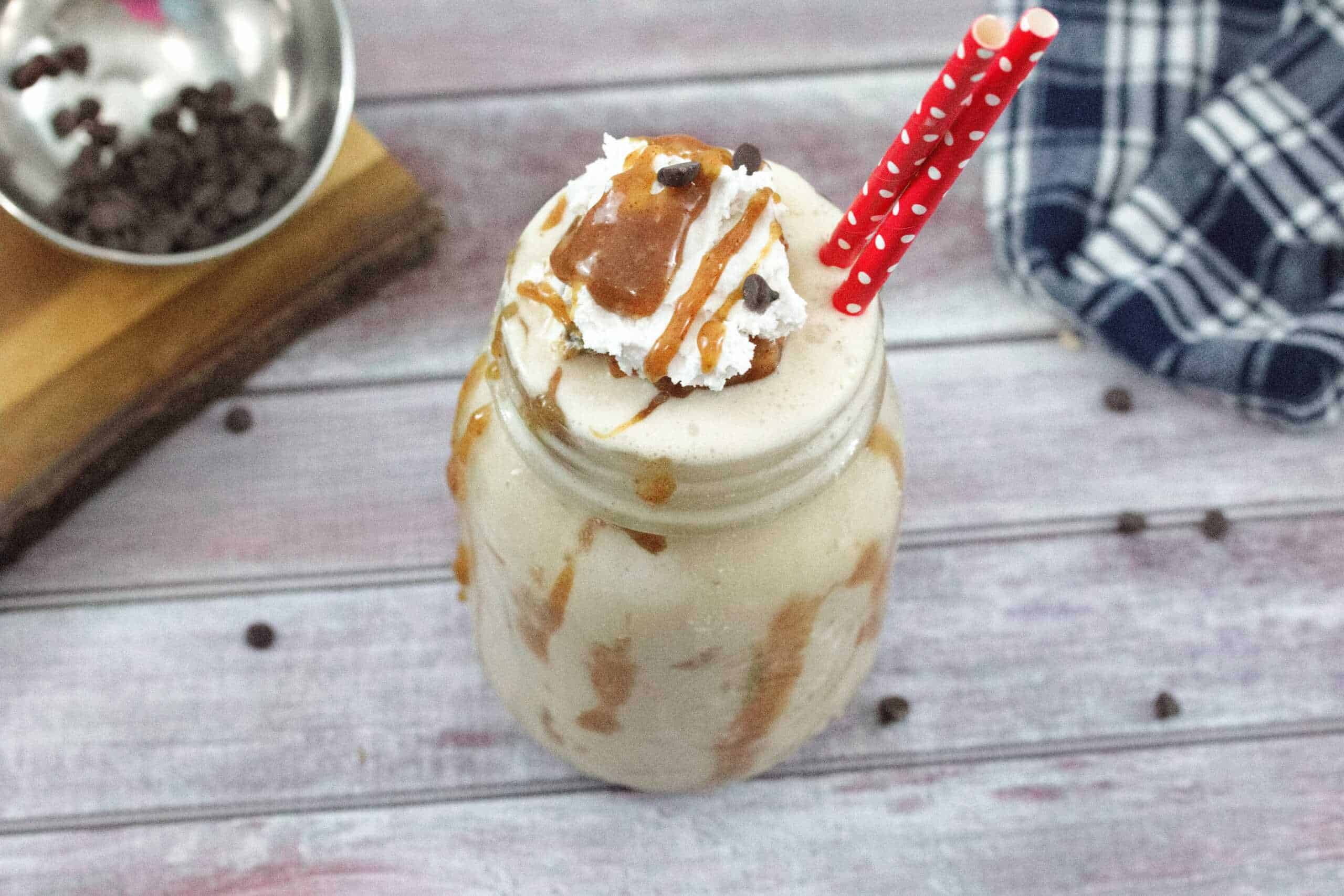 Mason jar with caramel protein shake topped with coconut whipped cream, caramel sauce and chocolate chips two red straws in the smoothie and chocolate chips on a cutting board behind it.