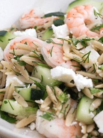 A shallow white bowl with orzo, shrimp, sliced cucumbers, and feta cheese.