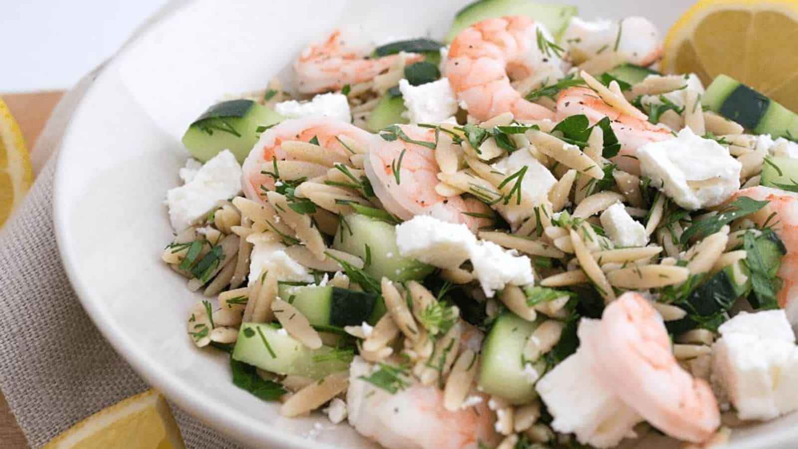 A shallow white bowl with orzo, shrimp, sliced cucumbers, and feta cheese.