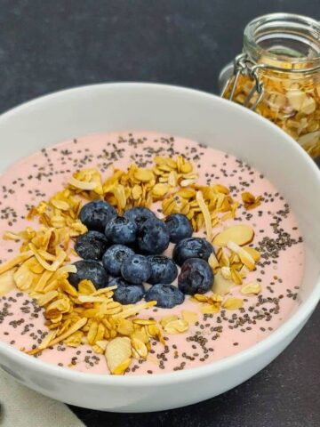 A bowl of pink yogurt topped with granola, chia seeds, and fresh blueberries, with extra blueberries and a jar of granola on the side.