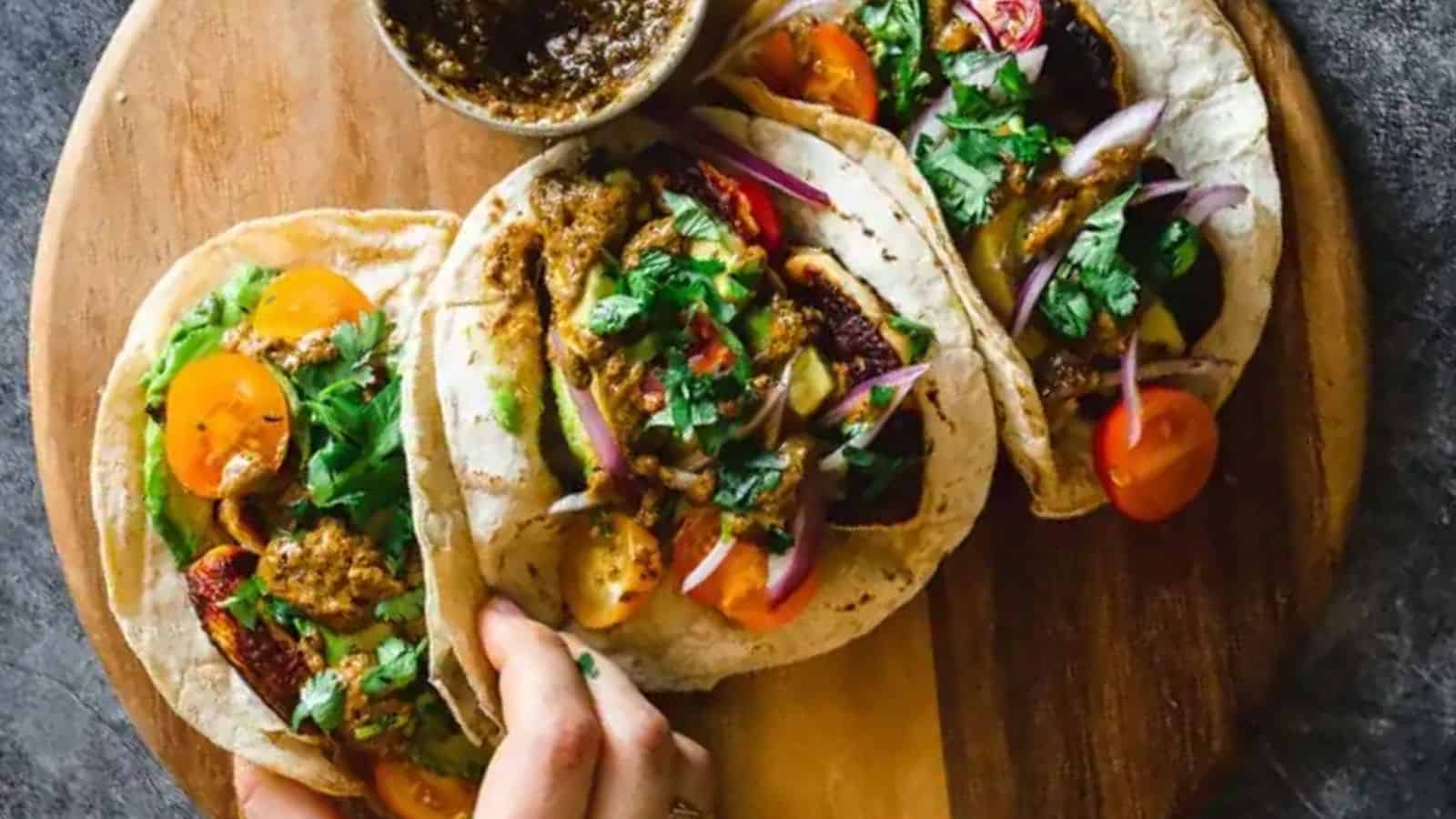 A hand grabbing a piece of halloumi ttacos with pepita salsa, cherry tomatoes, and avocados from a wooden board.