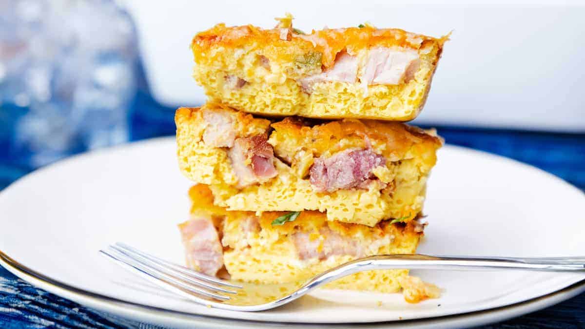 Three stacked slices of ham and egg casserole with melted cheese on a white plate, with a fork to the side.