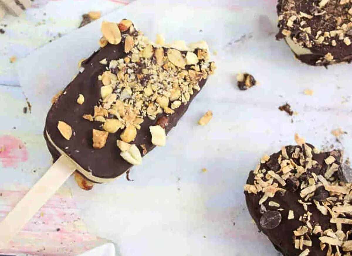 Homemade ice cream bar dipped in chocolate on a plate being covered in peanuts with more ice cream bars around it.