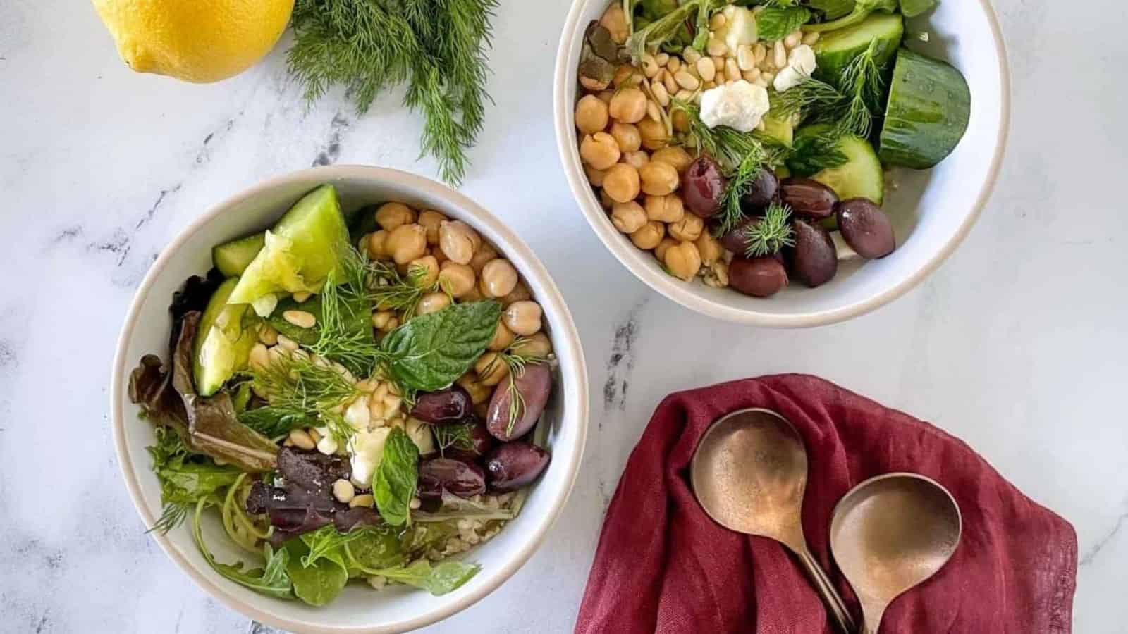 Two grain bowls are shown on a white marble counter with forks, a red linen, dill, and a lemon.