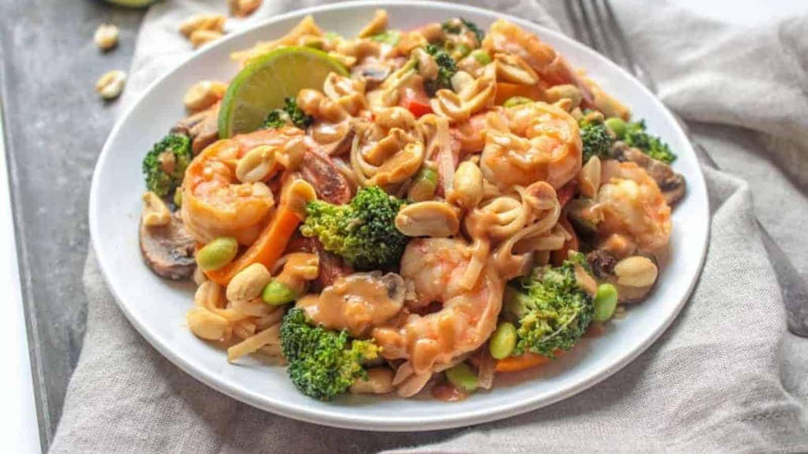 Shrimp, broccoli, bell peppers, mushrooms, and brown rice noodles drizzled with peanut sauce on a white plate.