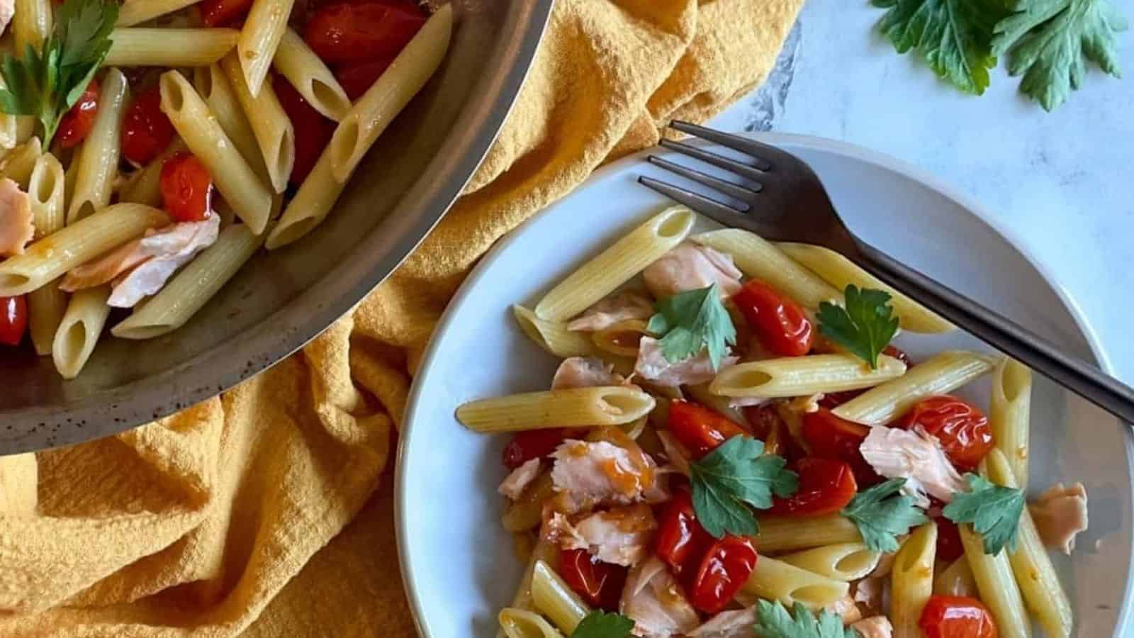 Penne pasta with burst cherry tomatoes, shallots, garlic, and parsley on white plate next to a pan filled with pasta.