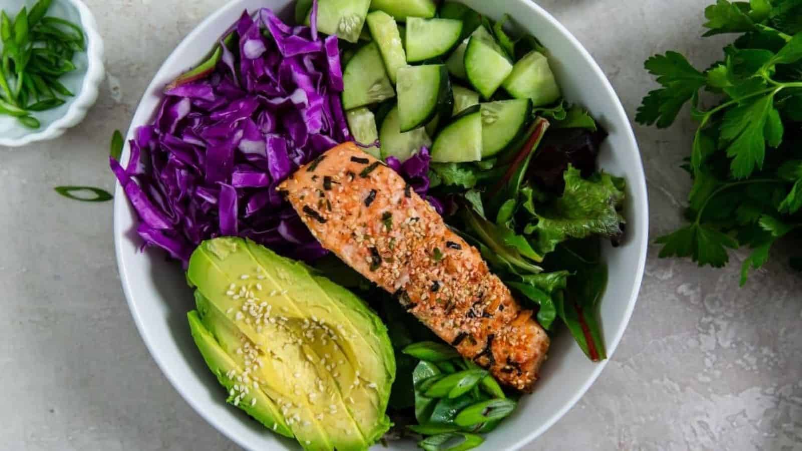 Sesame Salmon Bowl with cucumber, avocado, sesame seeds, red cabbage, green onions and spring mix in a white bowl.