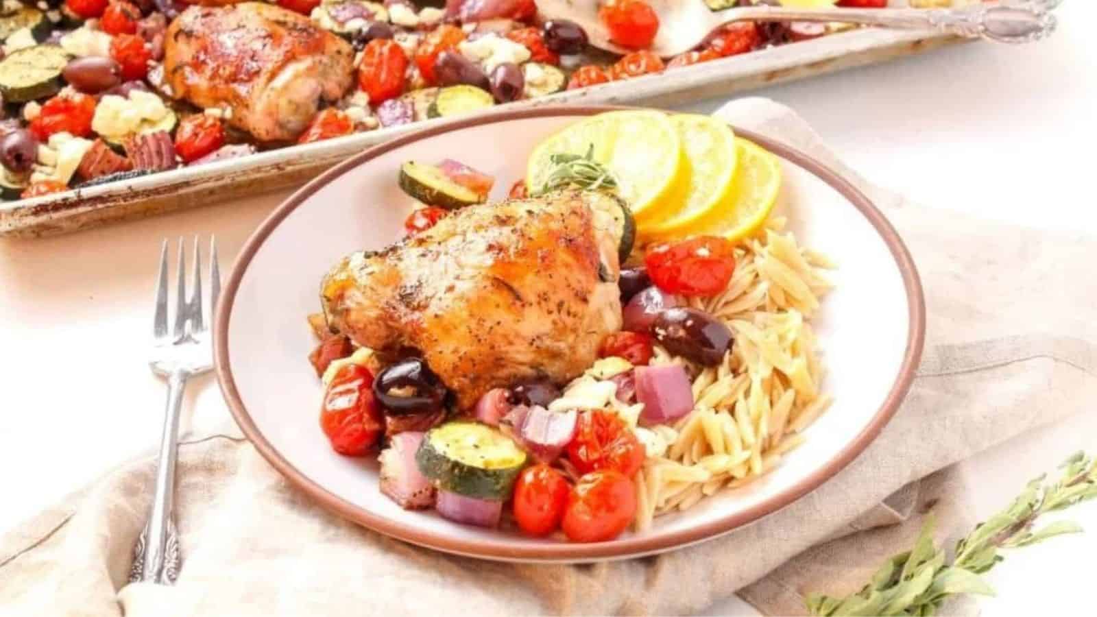 A shallow white bowl filled with a roasted chicken thigh, roasted zucchini, cherry tomatoes, red onion, kalamata olives, feta cheese, and orzo.