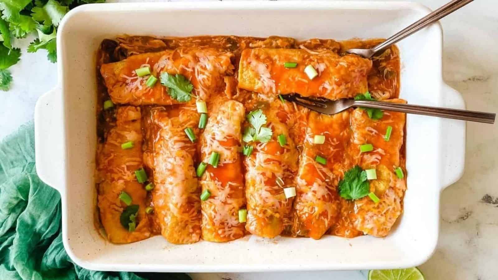 A baking dish full of chicken enchiladas is surrounded by cut limes, a halved avocado, and a green linen.