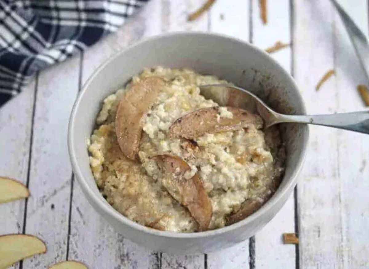 Bowl of protein oatmeal with apples on top and a blue linen behind it and a spoon dipping into the oatmeal.