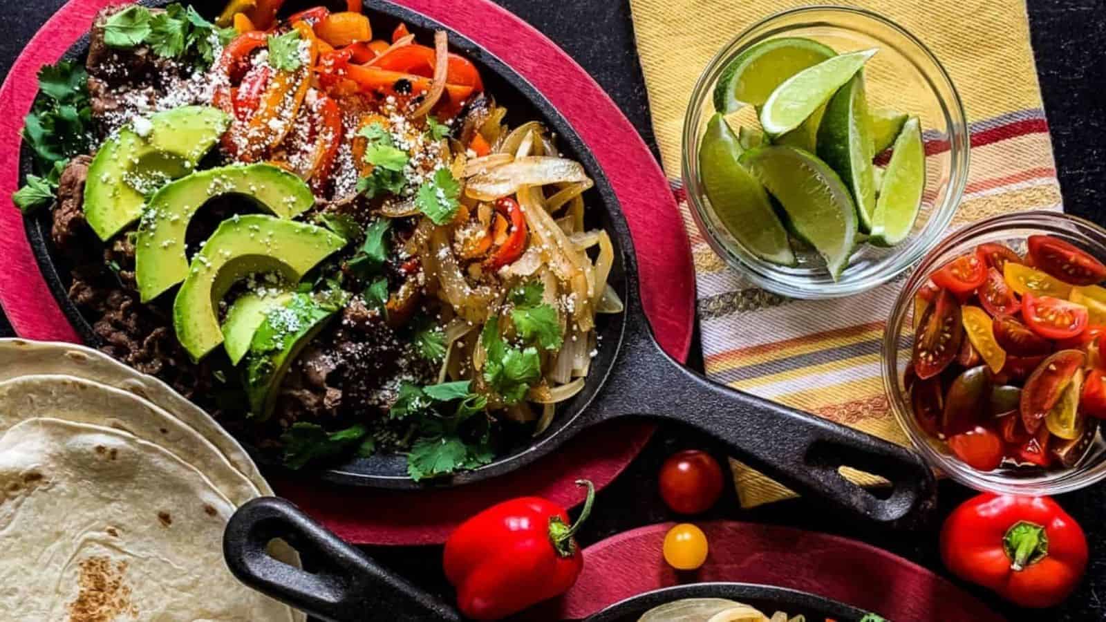 A cast iron skillet with hot beef fajitas and seared vegetables alongside bowls of lime wedges, tomatoes, and tortillas.