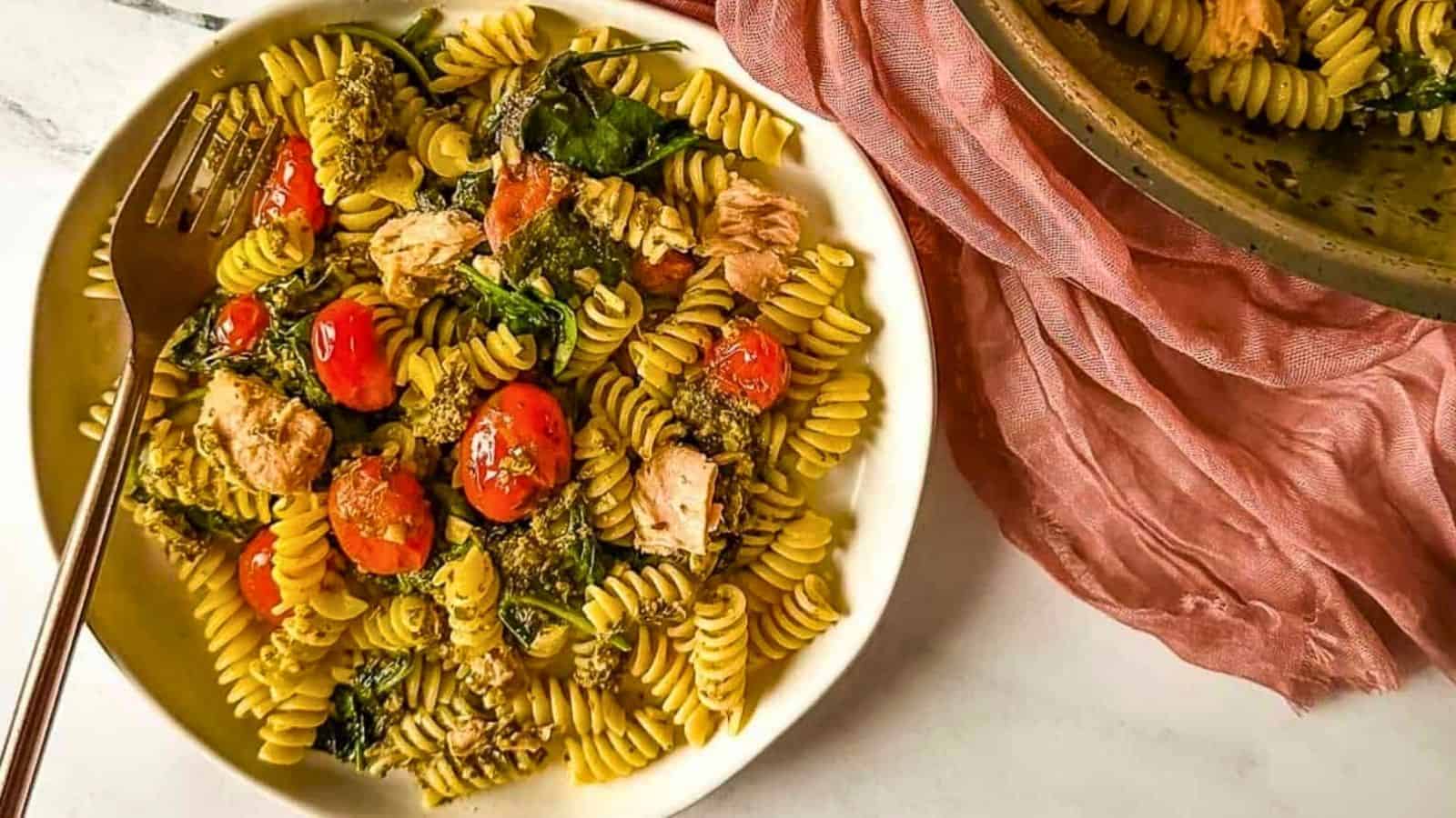 A plate of tuna pesto pasta with cherry tomatoes, a copper fork, and a pink linen next to a pan full of tuna pesto pasta.