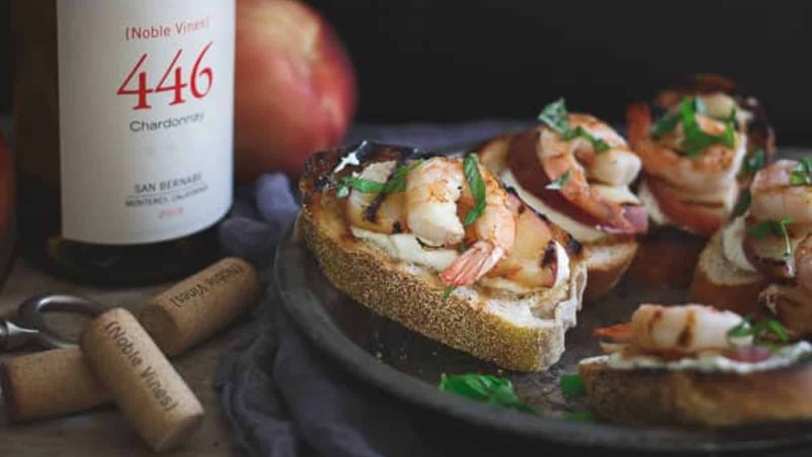 A close-up photo of Whipped Lemon Burrata Crostini with Grilled Shrimp and Peaches on a plate with a bottle of chardonnay.