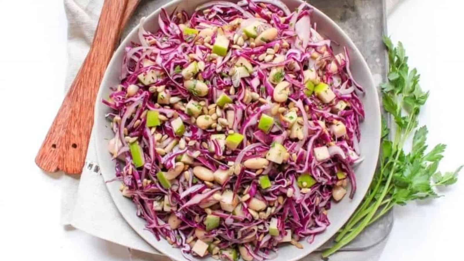 A white plate with cabbage slaw, sliced apples, and white beans on a metal sheet pan with a beige linen napkin.