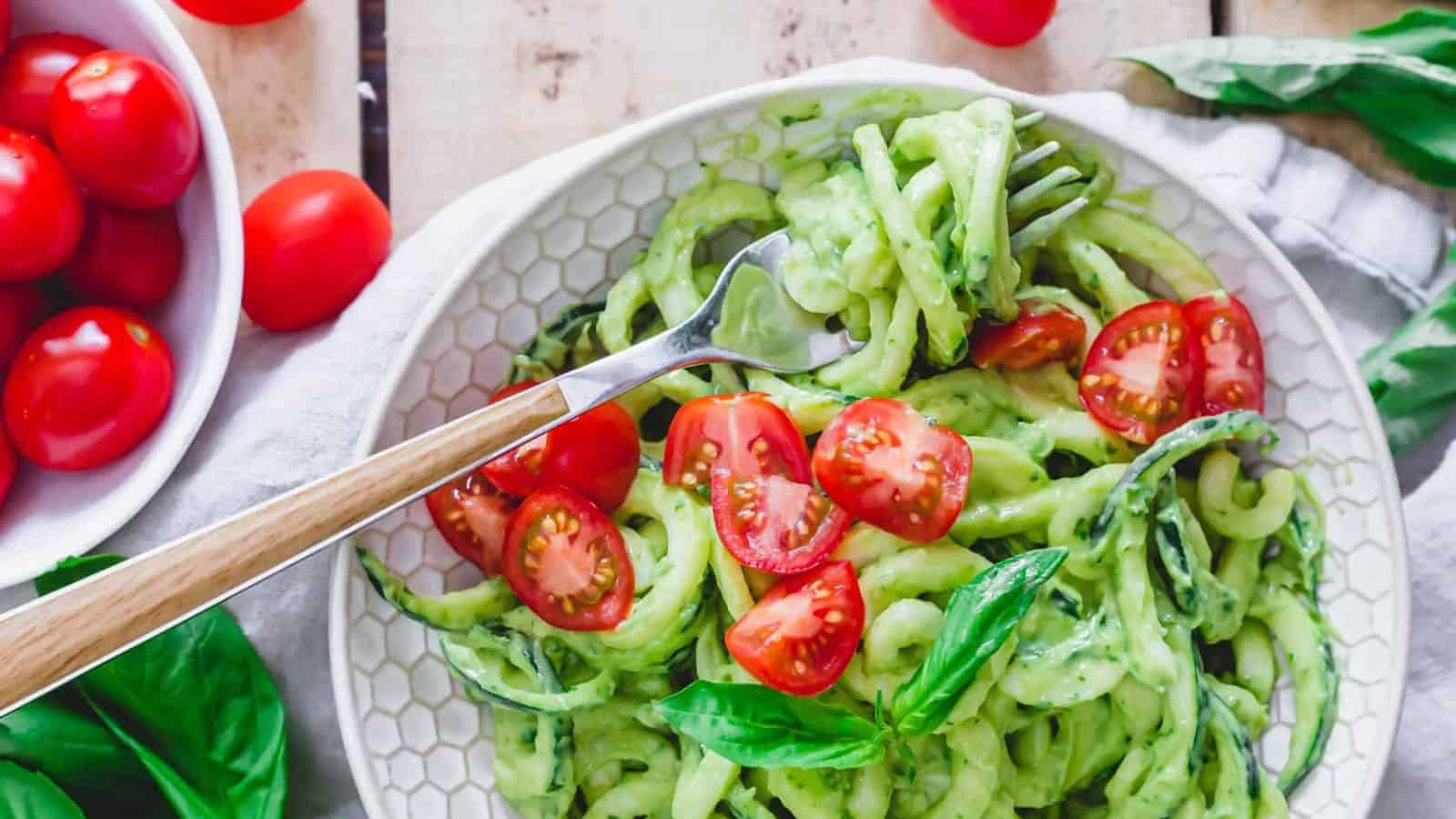 A plate of zucchini noodles with creamy avocado sauce, fresh tomatoes on top and a fork.