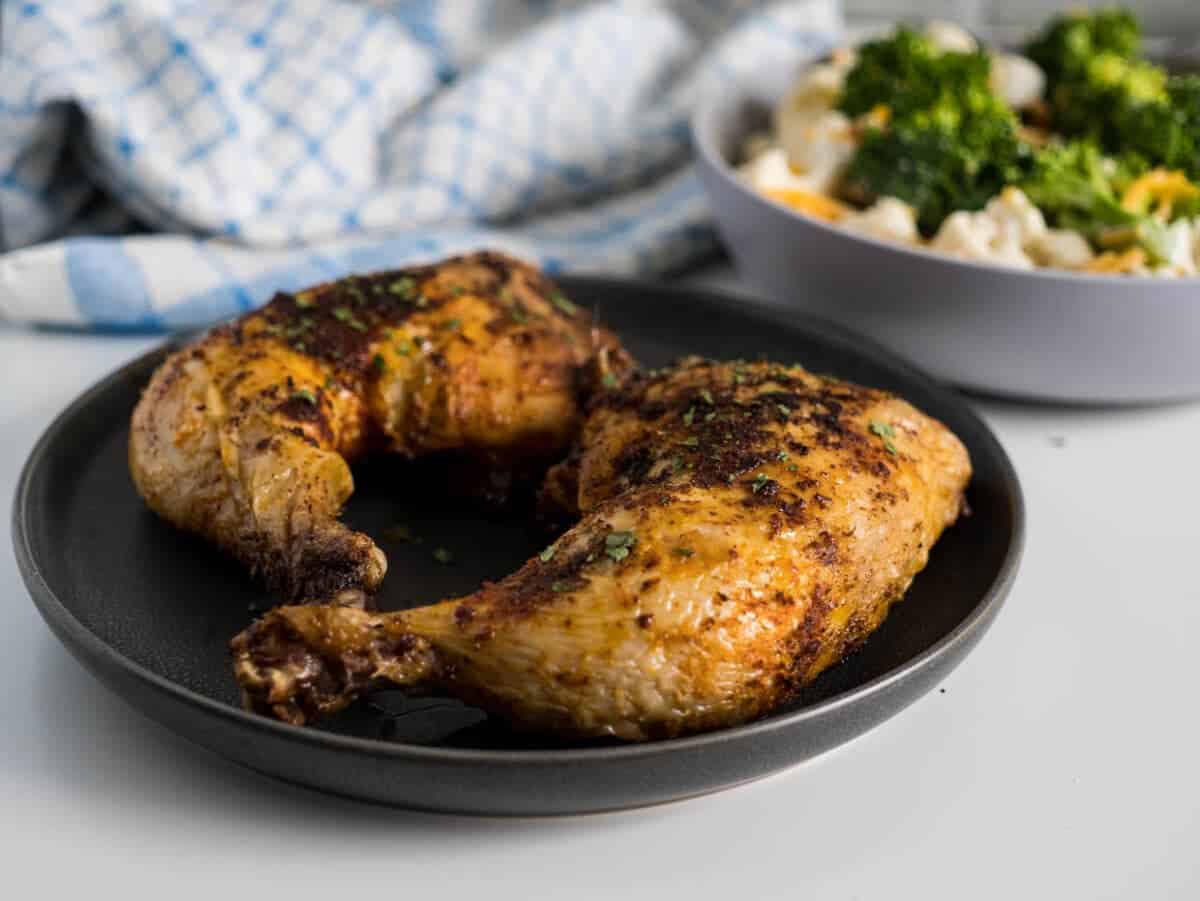 Two chicken leg quarters on a grey plate sprinkled with parsley with a broccoli salad in the background.