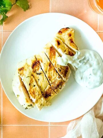 Grilled chicken with cilantro crema on a white plate surrounded by limes, cilantro, and a glass of beer.
