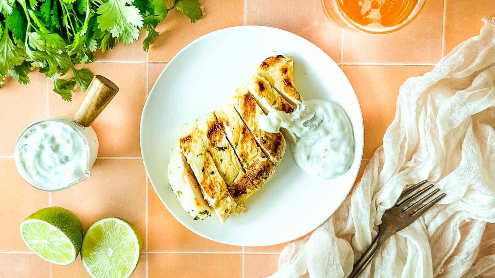 Grilled chicken with cilantro crema on a white plate surrounded by limes, cilantro, and a glass of beer.