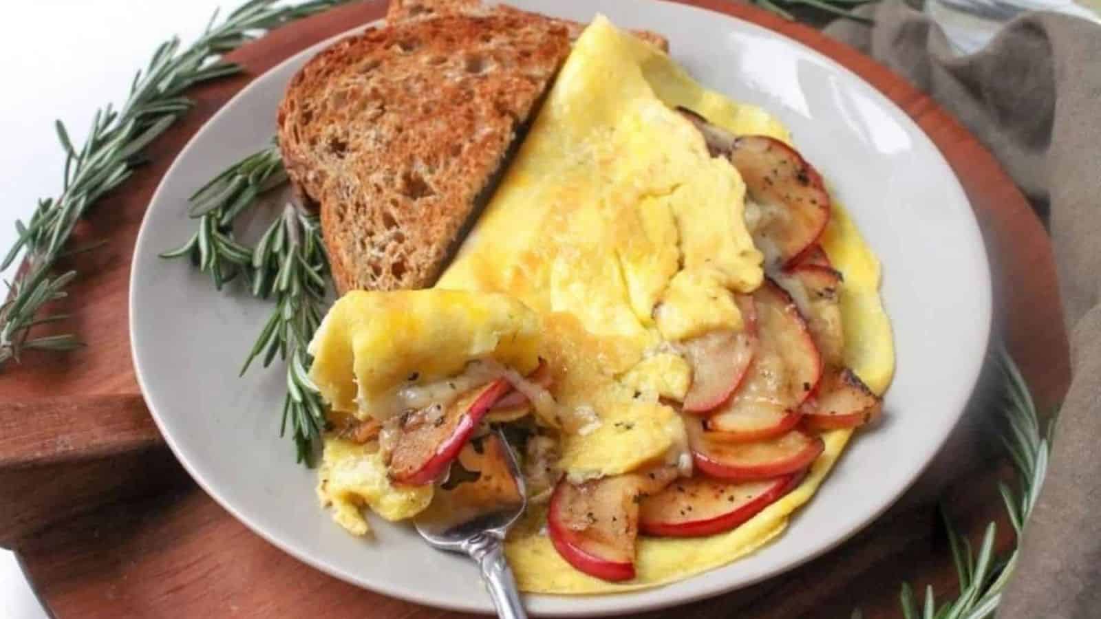 An apple white cheddar omelette with a slice of toast and a fork on a plate sitting on a wooden platter with fresh herbs surrounding it.