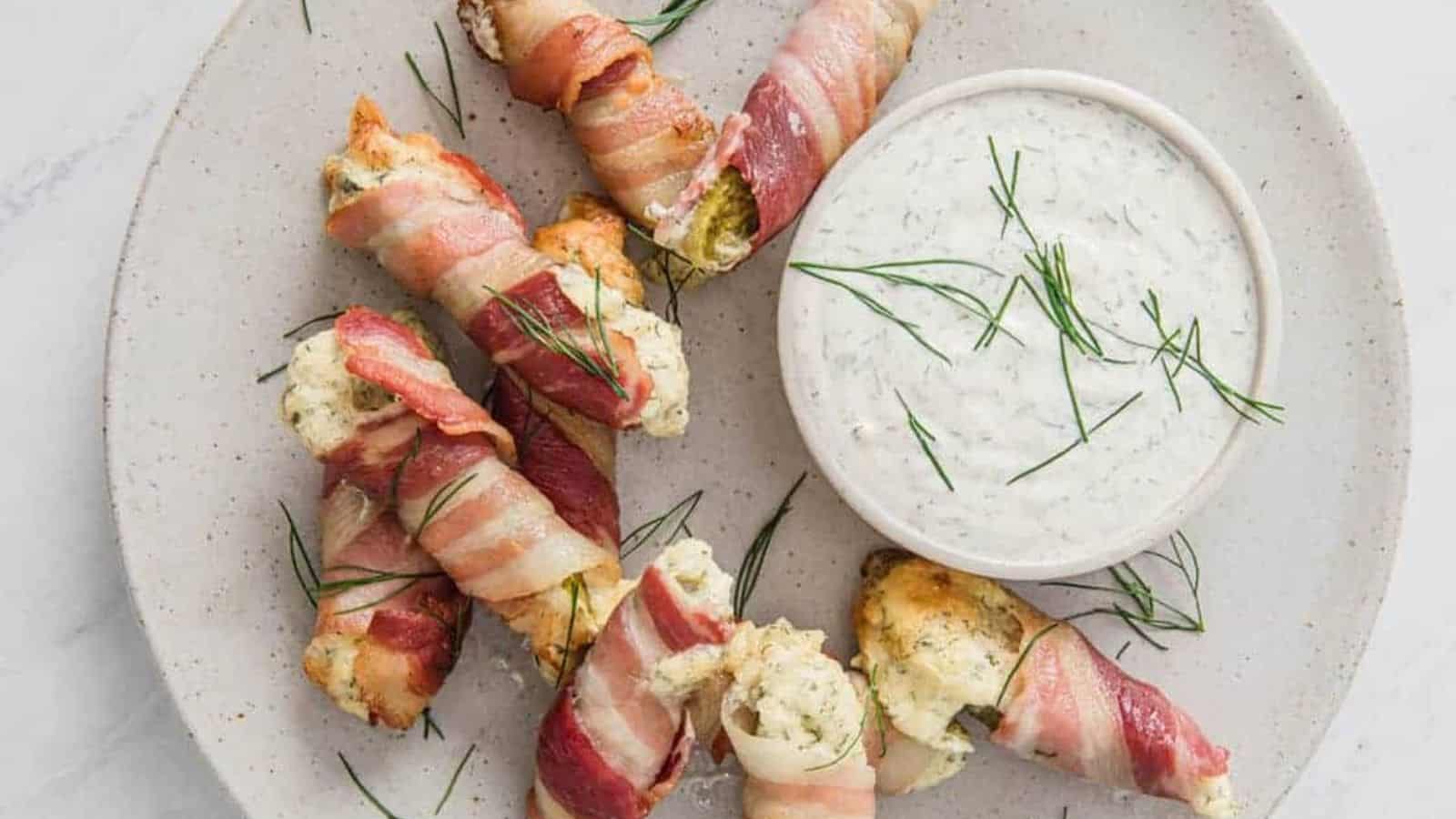 Bacon pickle boats stuffed with cream cheese on a plate next to a bowl of dip.