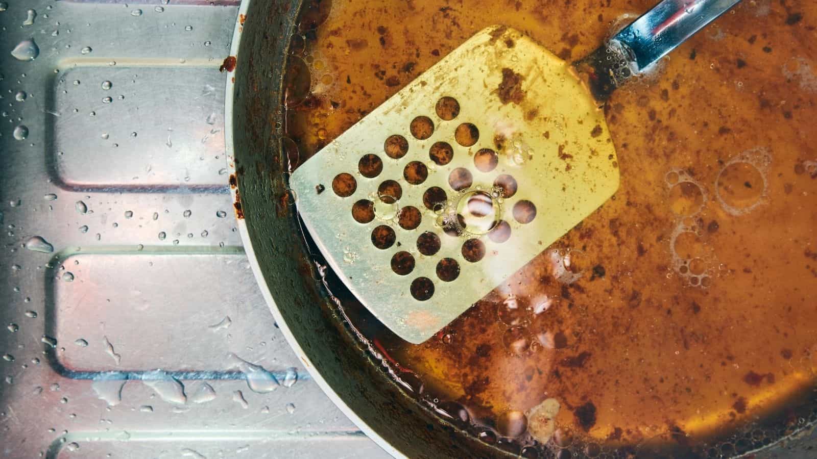 Greasy steel pan and perforated spatula on steel sink with water droplets.