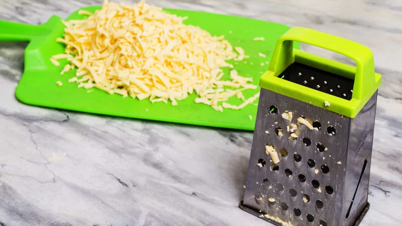 Box grater with grated cheese on a green cutting board.