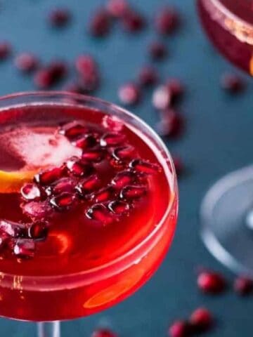 Two champagne pomegranate cocktails in coupe glasses on a dark blue background scattered with pomegranate seeds.