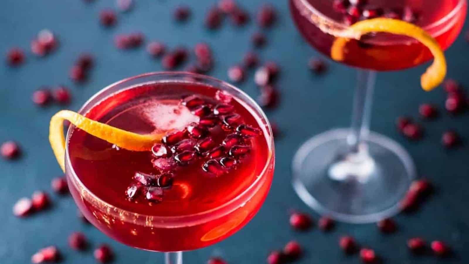 Two champagne pomegranate cocktails in coupe glasses on a dark blue background scattered with pomegranate seeds.