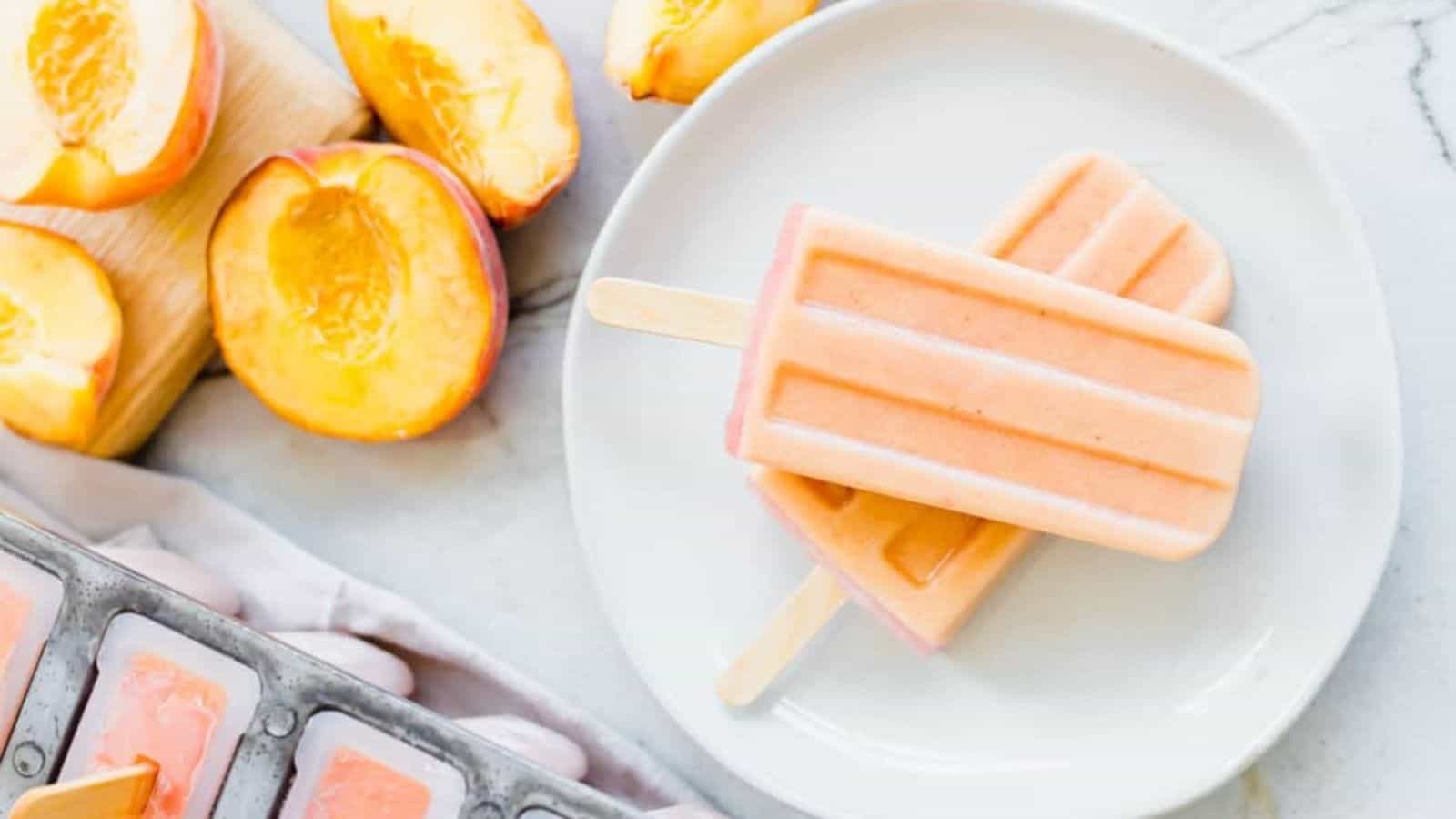 Top view image of 2 creamy peach popsicles on a plate with slices of peach in the background.