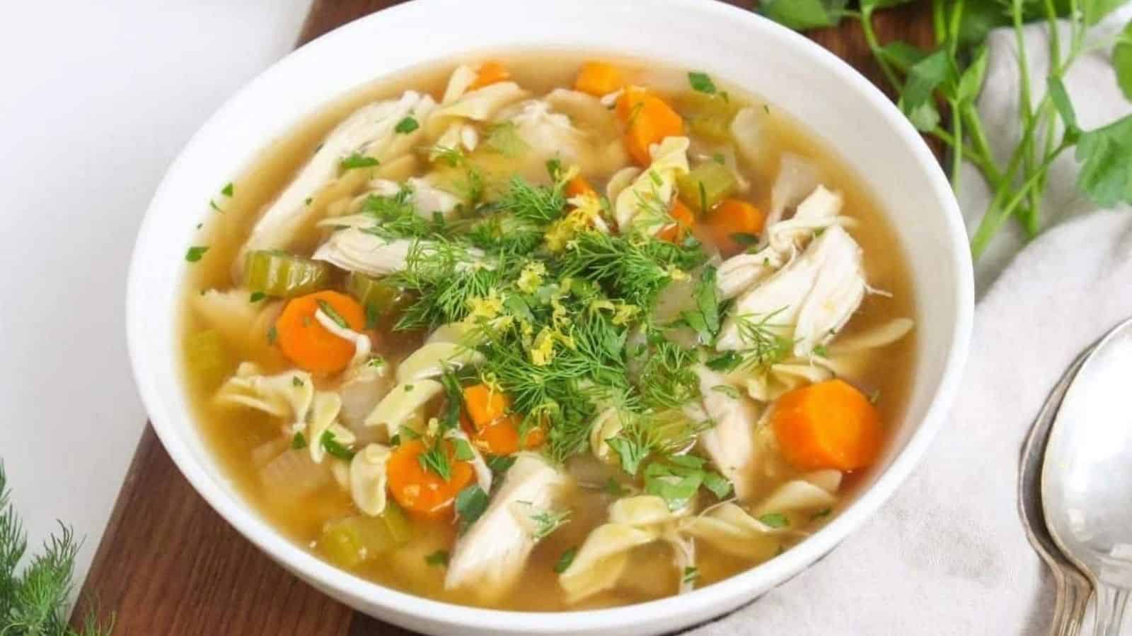 A white bowl with chicken noodle soup topped with fresh herbs.