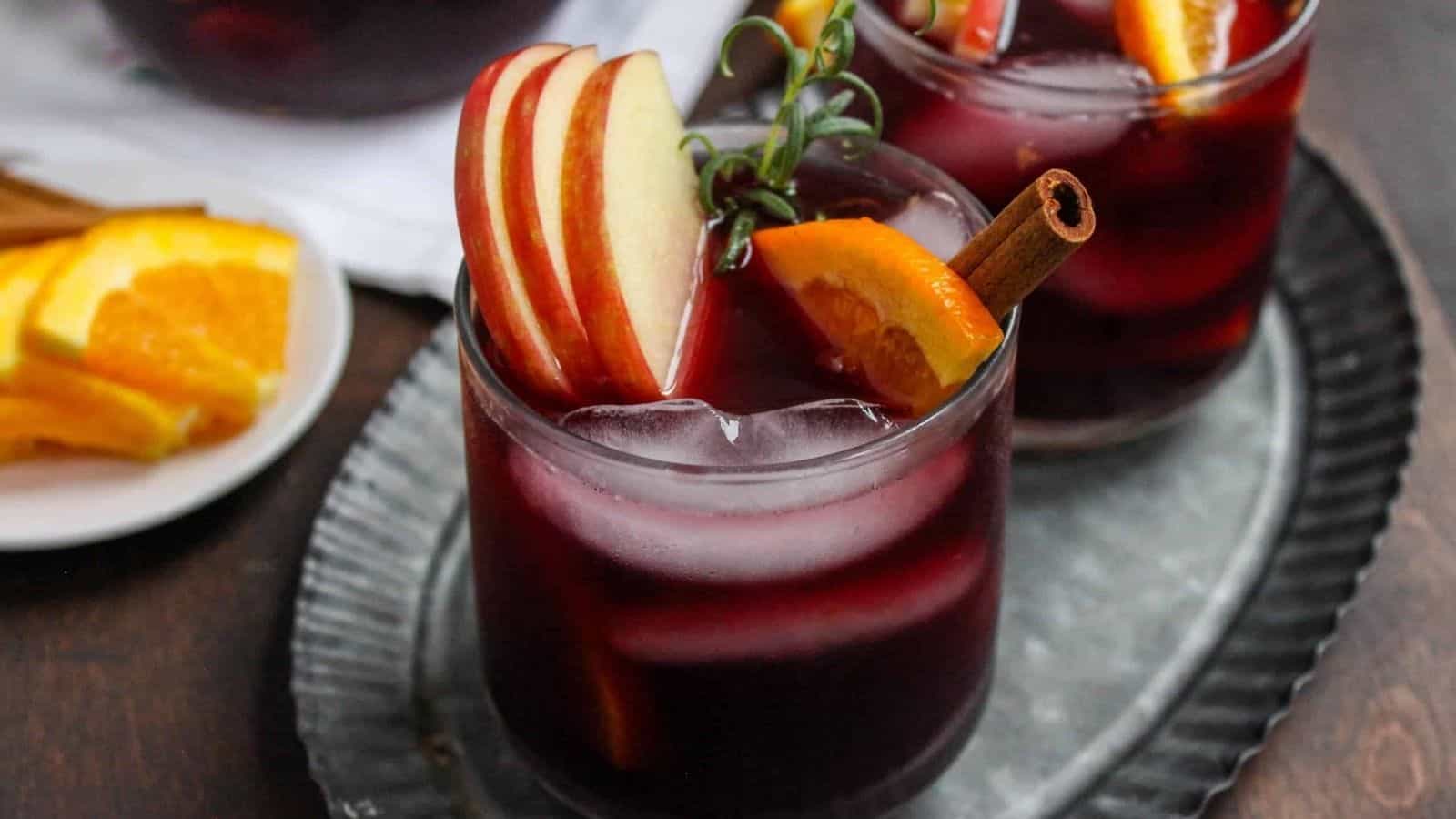 A glass of red wine bourbon sangria with apple slices and cinnamon sticks sitting on a metal plate.