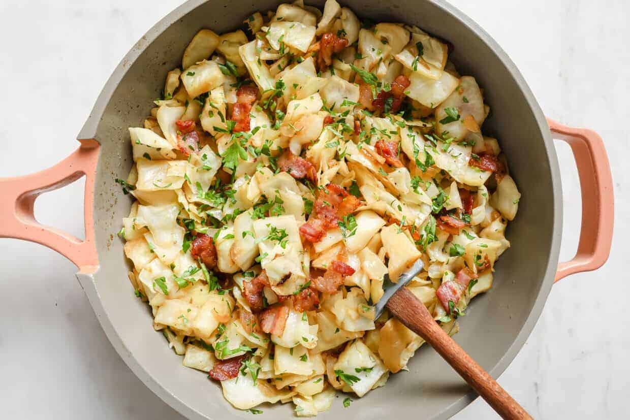 A pan full of bacon and cabbage with a wooden spoon.