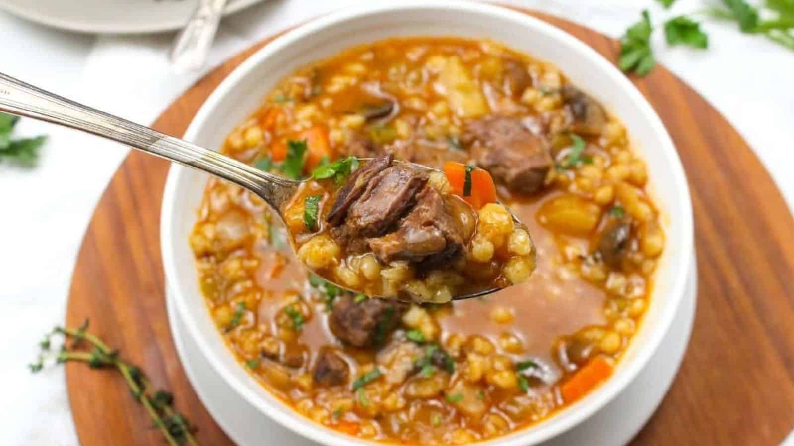 A spoonful of beef and barley soup with a bowl in the background.