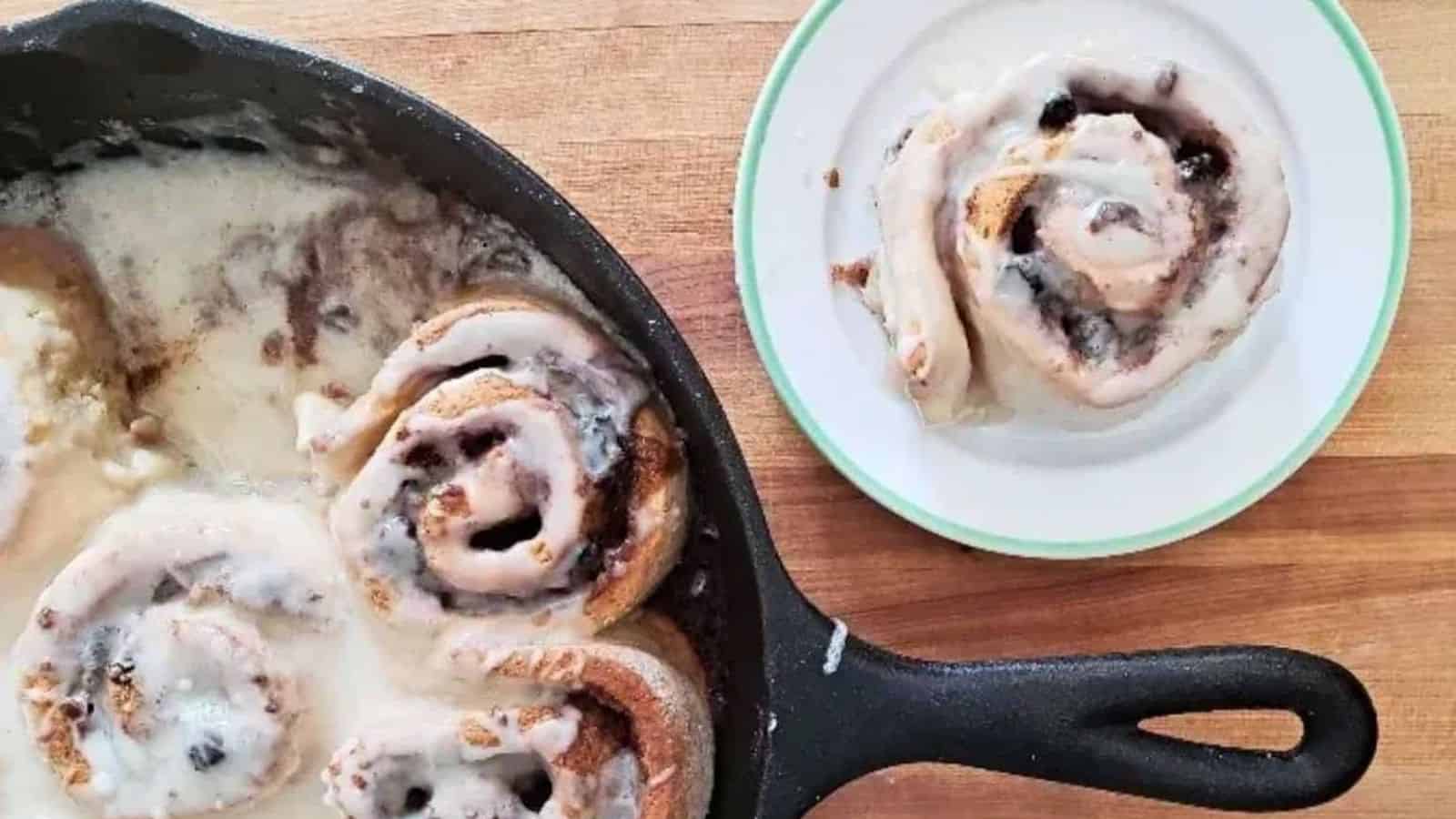 Image shows an overhead shot of Cinnamon rolls in a skillet on a wooden table with one cinnamon roll on a plate.