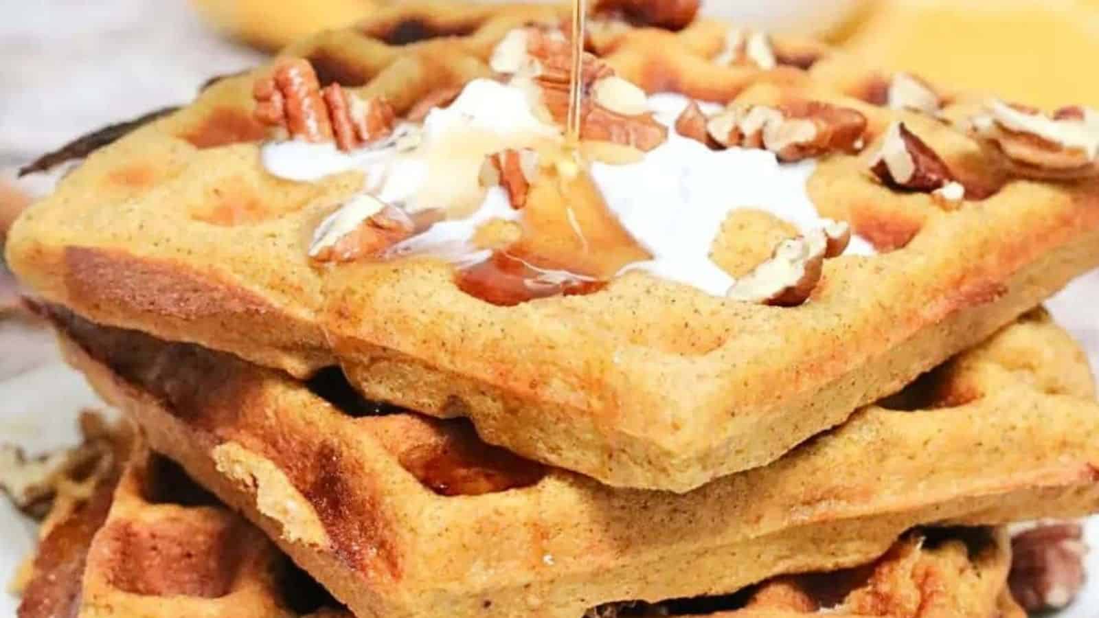 three gluten-free pumpkin waffles stacked on a white plate with coconut cream, maple syrup and pecans on top. A waffle maker beside the plate and a bowl of pecans and syrup behind it