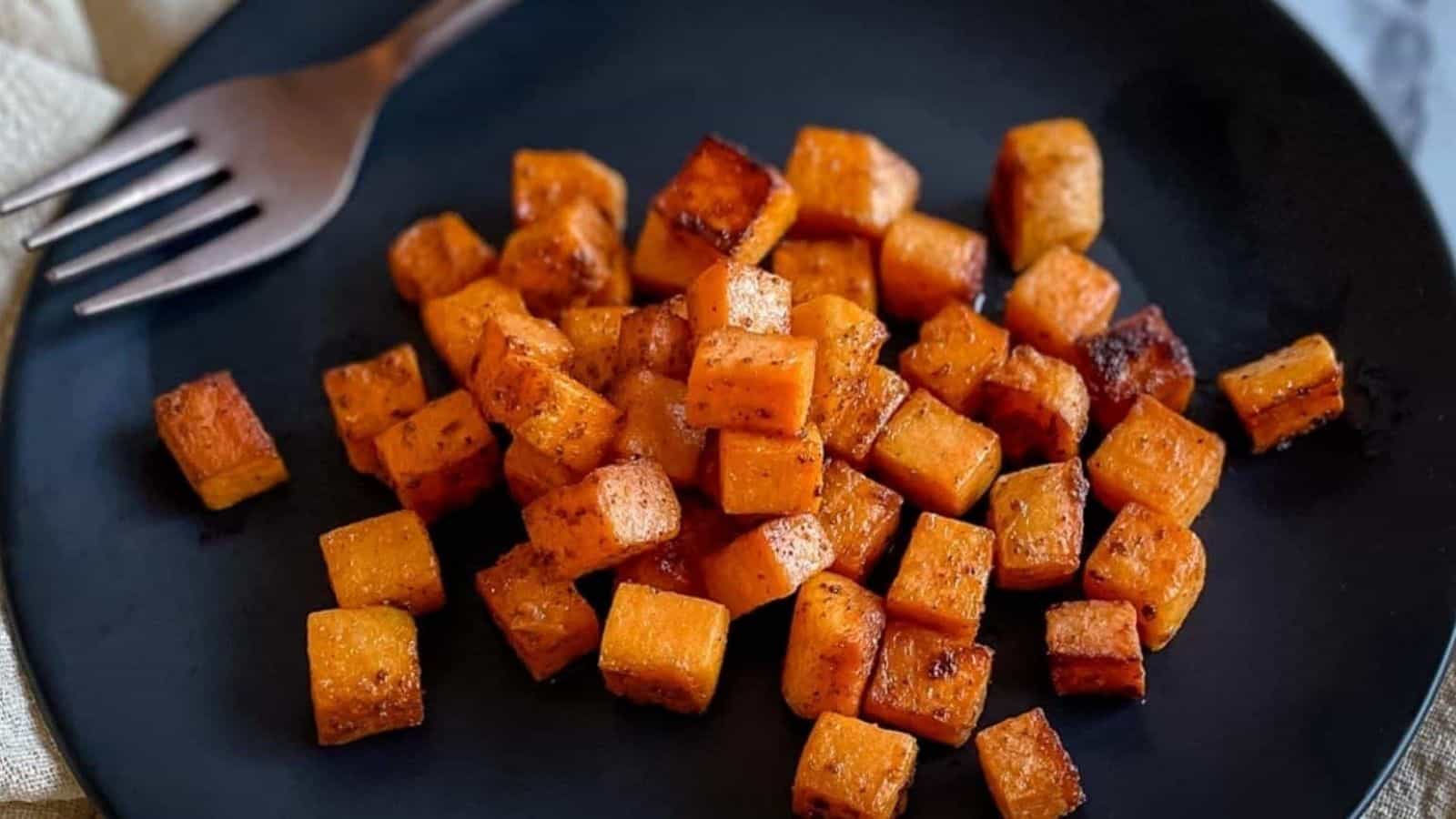 Sautéed sweet potatoes are shown on a black plate with the tines of a fork and a cream-colored linen.