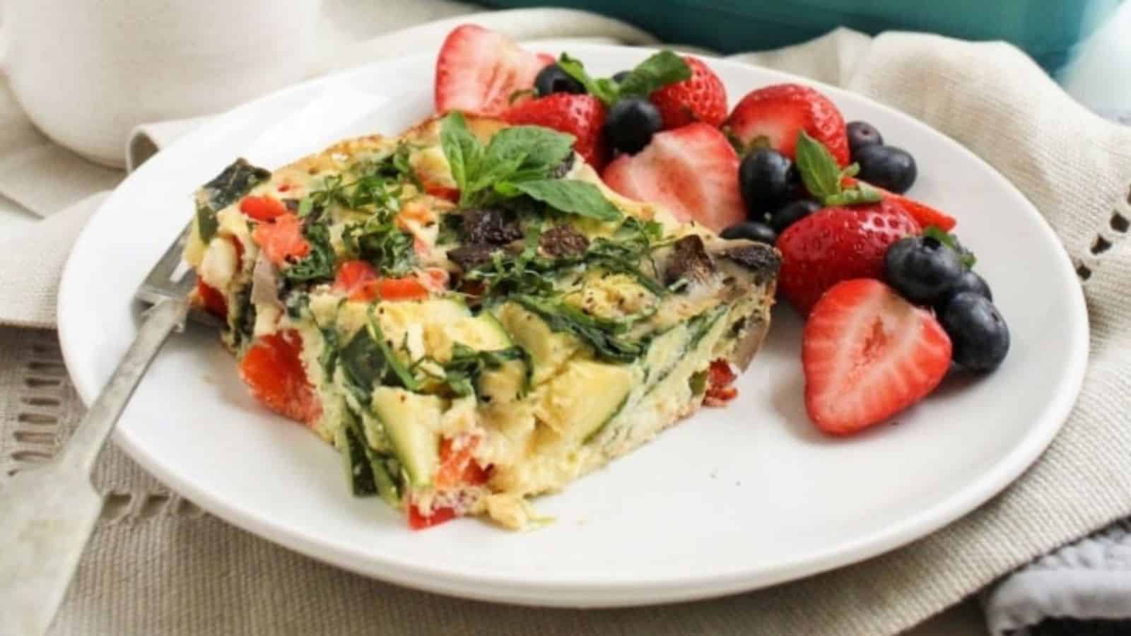 A slice of smoked salmon egg casserole on a white plate with strawberries and blueberries, with a mug of coffee behind it.