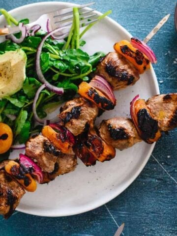 A plate of Smoky Grilled Lamb Kebabs with Apricots and a side of greens with a slice of avocado.
