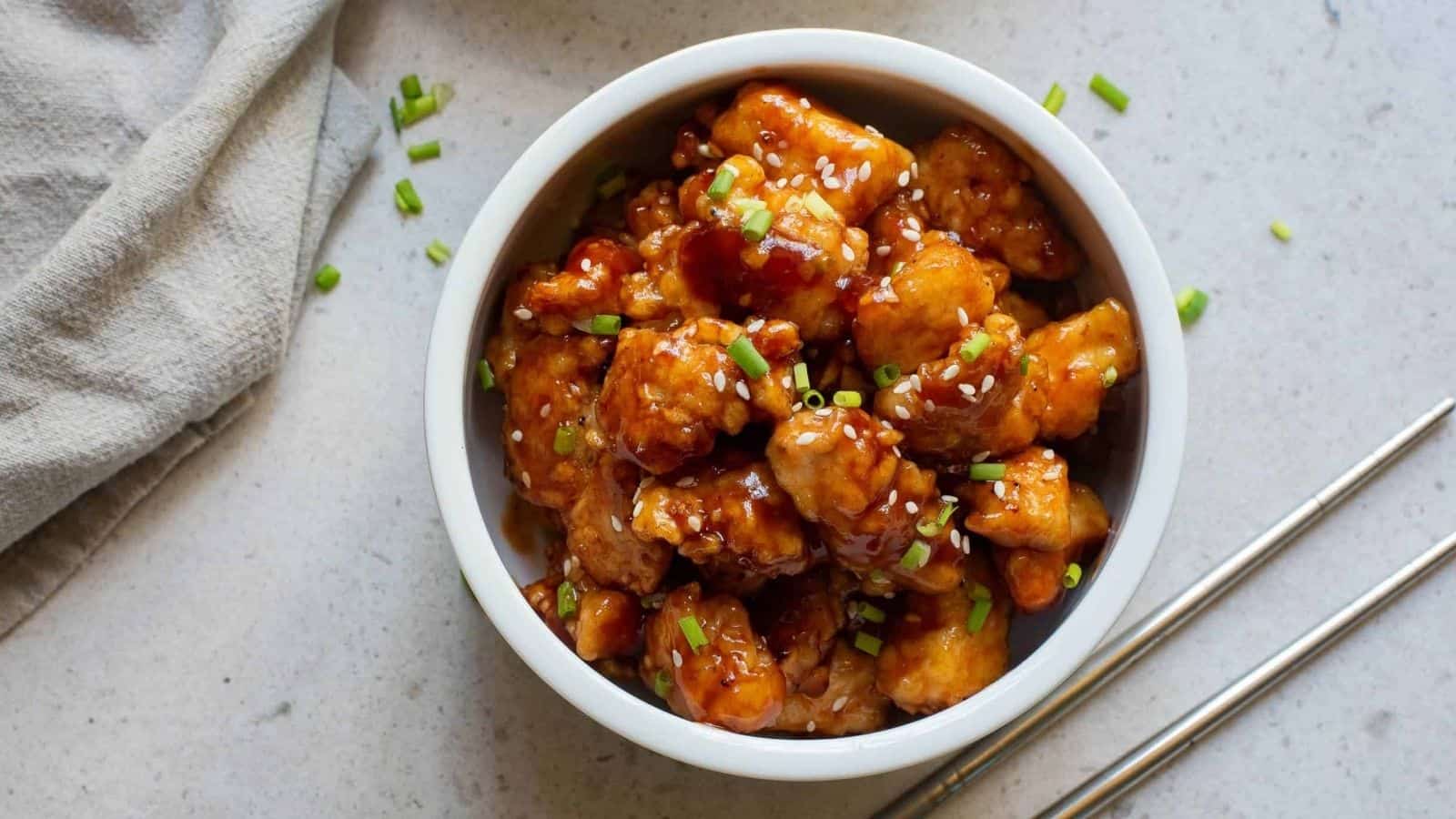 A bowl of asian chicken in a white bowl with chopsticks.