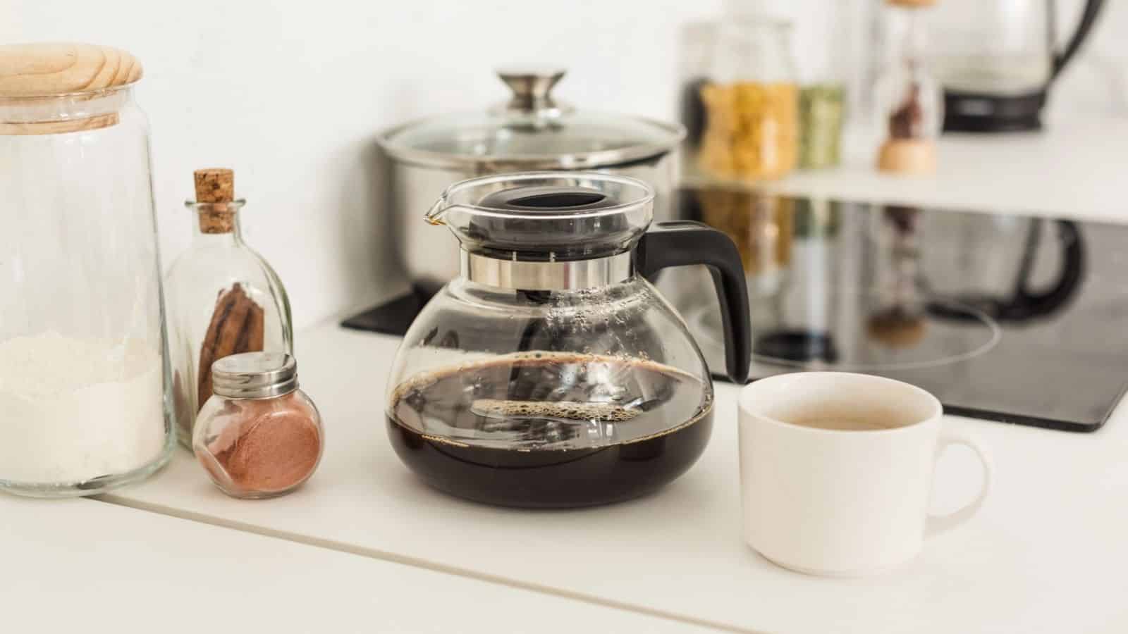 Close up view of coffee maker, cup and glass jars near stove on tabletop at kitchen.