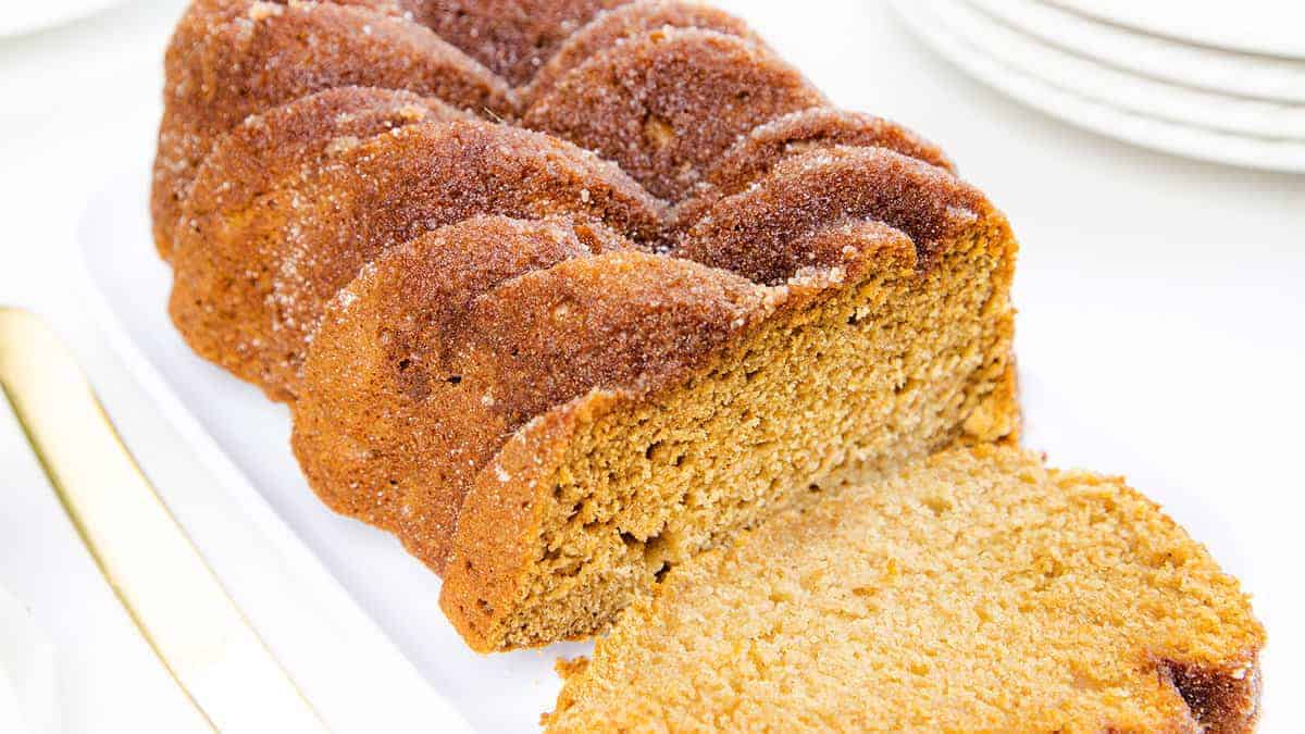 The image shows a loaf of applesauce bread, golden-brown with a sugary crust, partially sliced on a white plate next to a gold-colored.