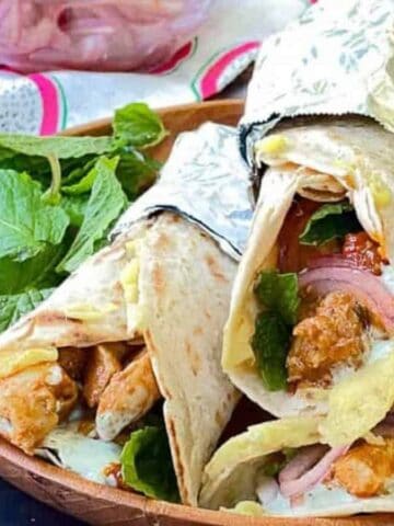 Low angle shot of 3 chicken kathi rolls wrappedin foil on a wooden plate with fresh mint leaves.