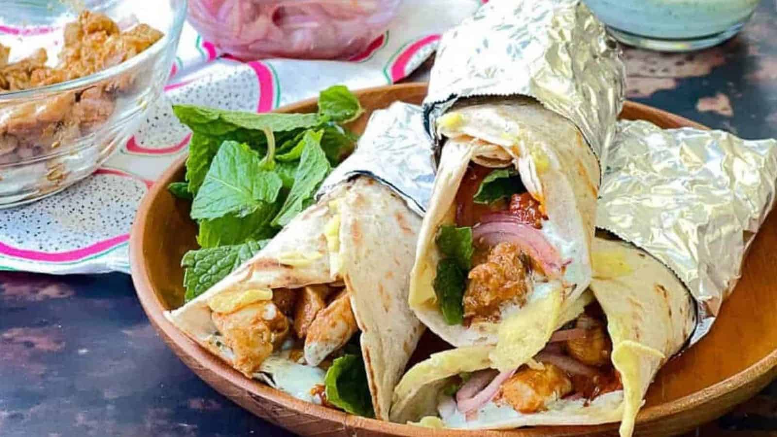 Low angle shot of 3 chicken kathi rolls wrappedin foil on a wooden plate with fresh mint leaves.