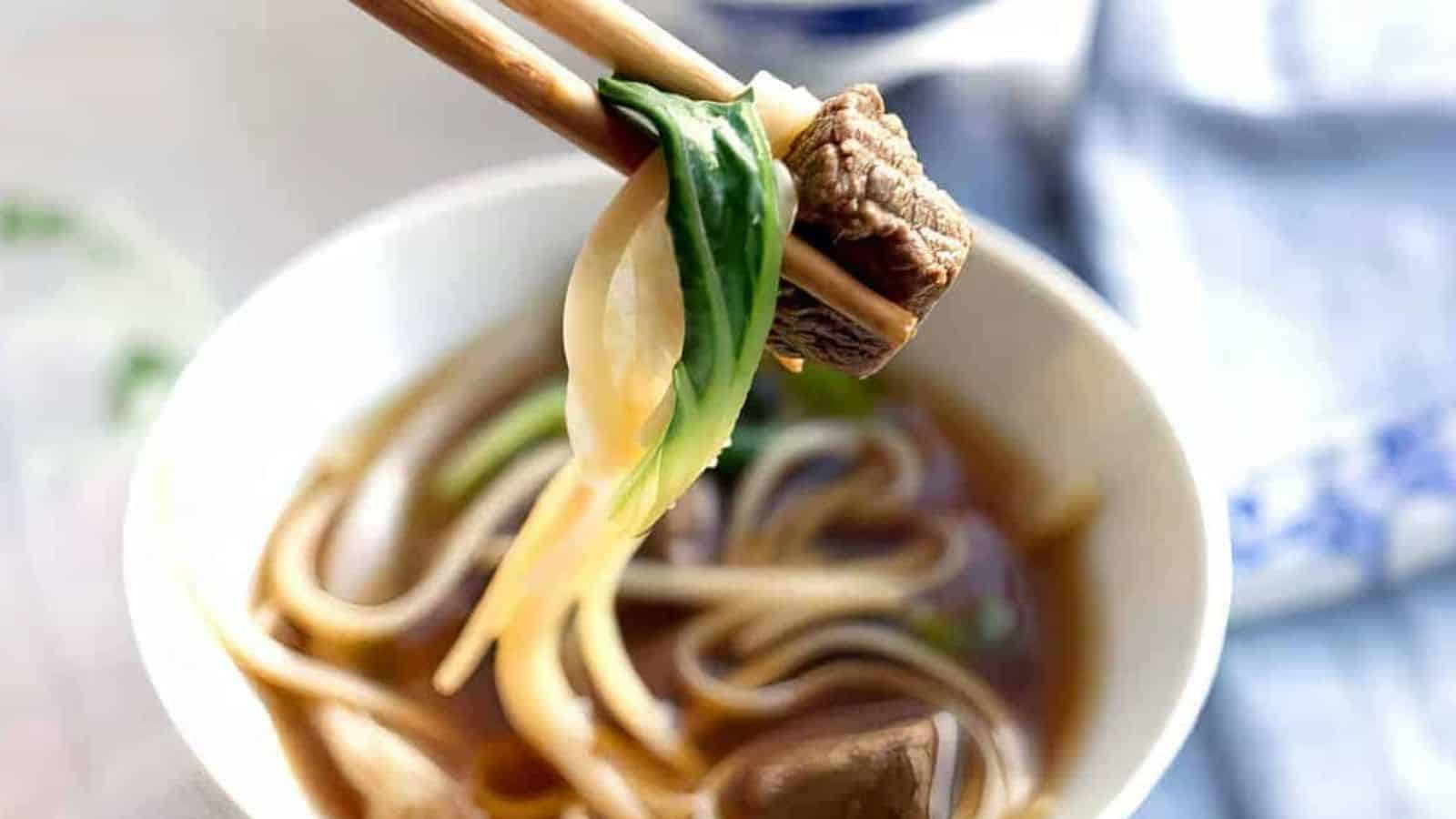 A white bowl containing beef noodle soup with a dark broth, slices of beef, white noodles, and green scallions on top.