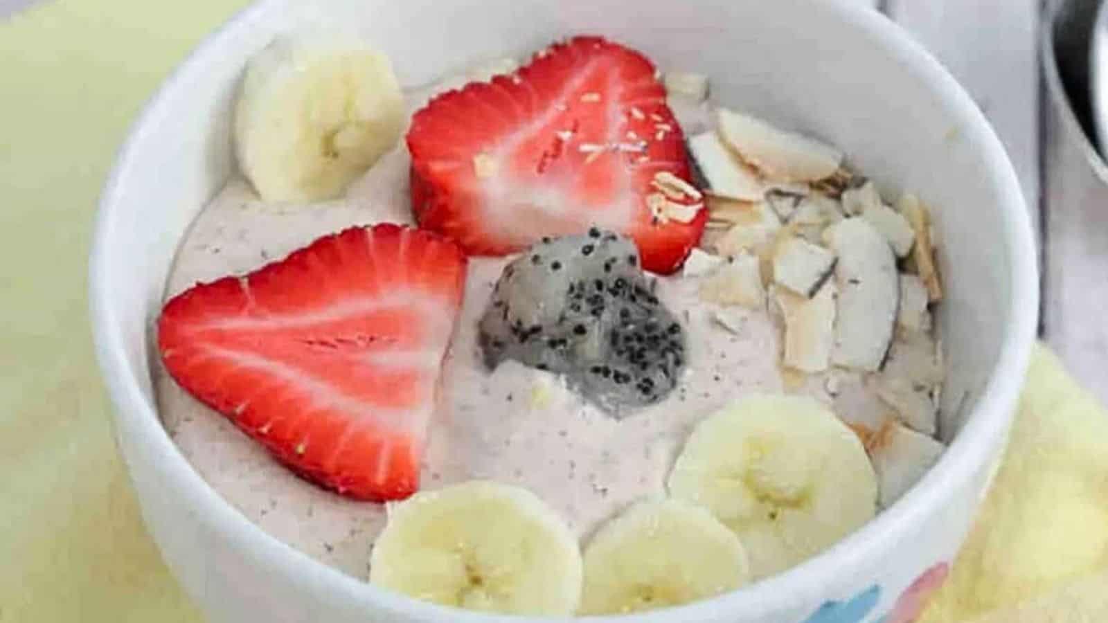 White bowl with a dragon fruit smoothie bowl in it topped with bananas, strawberries, and coconut on a yellow linen with berries, dragon fruit, and spoons around it.