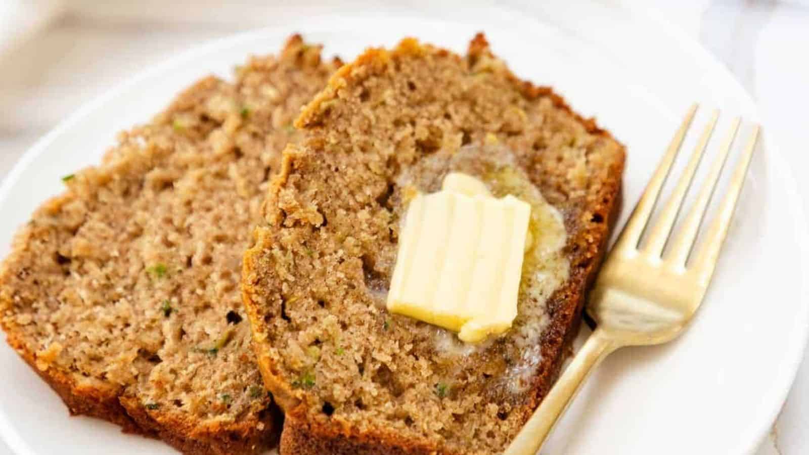 The image displays slices of moist zucchini bread with butter on a plate, alongside a fork.