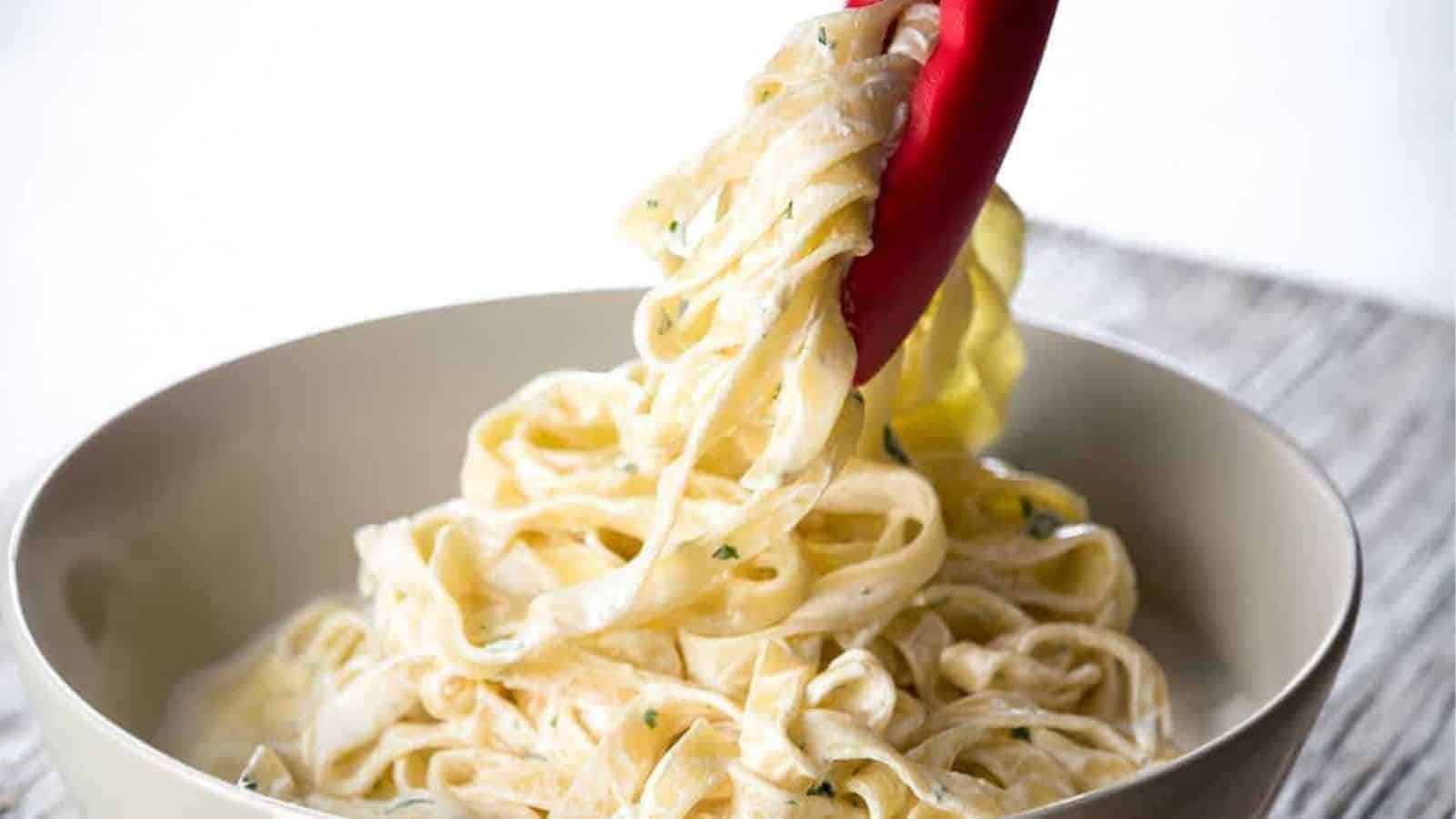 A close up image of pair of cooking tongs serving a portion of Fettuccine Alfredo into a bowl.